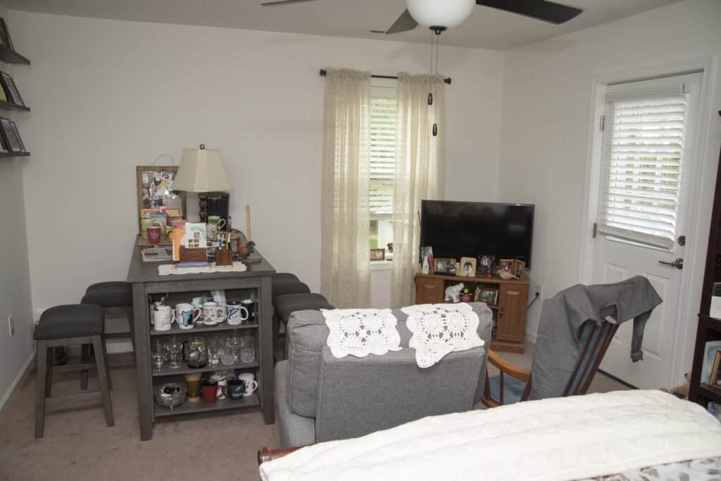 A cozy living room with gray armchairs, a TV on a wooden stand, a dining table with stools, shelves with mugs, and light curtains on windows. A ceiling fan is visible above, and decor items are arranged on surfaces.