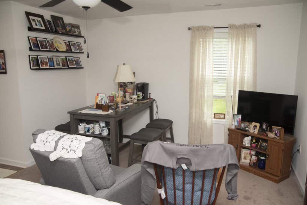 A cozy living room with a gray armchair, a wooden rocking chair, a small table with two stools, a TV on a wooden stand, and family photos on shelves and surfaces. Light curtains cover a window letting in daylight.