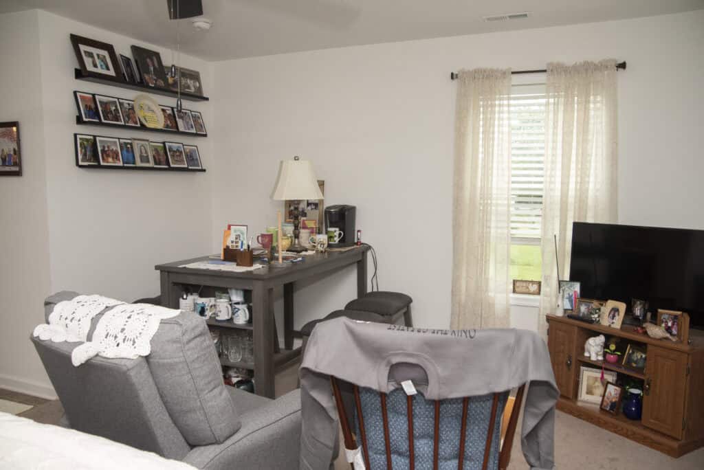 A cozy living room with gray chairs, a wooden table with a lamp, shelves of framed photos on the wall, a TV on a wooden stand, and a window with sheer curtains letting in natural light.