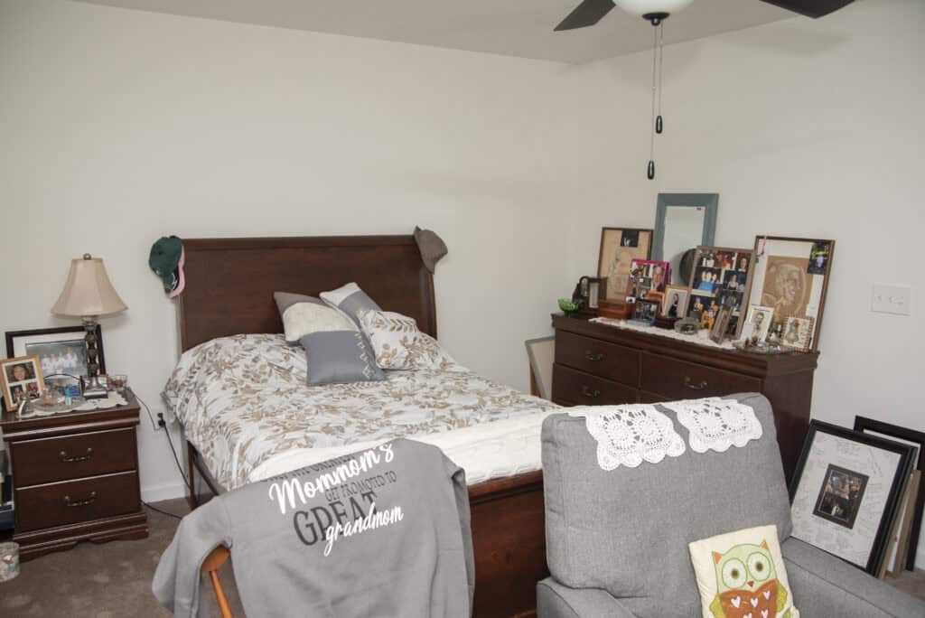 A cozy bedroom with a wooden bed, floral bedding, and many family photos on a dresser and nightstand. A gray chair with an owl pillow sits nearby, and a ceiling fan hangs above.