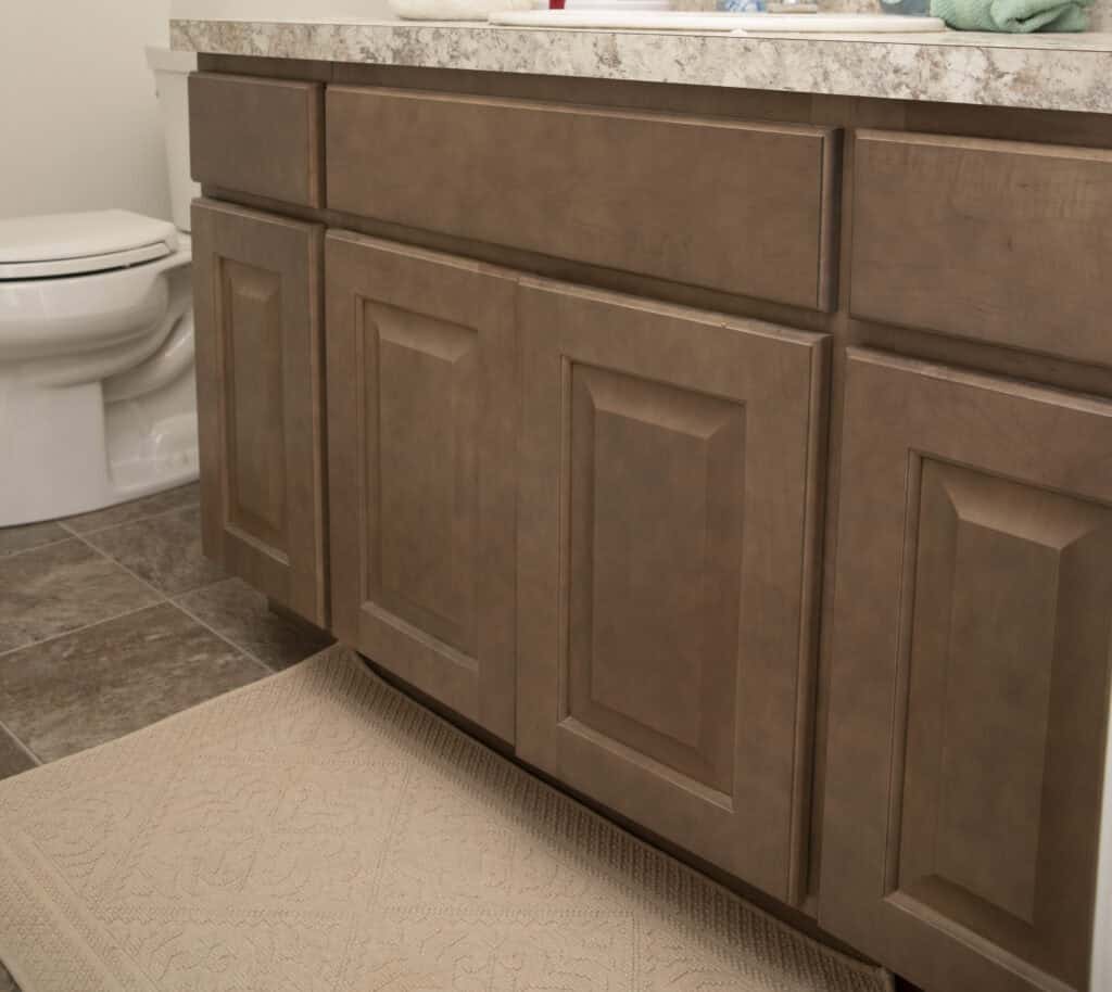 Bathroom with brown wooden cabinets, a beige patterned rug on a tiled floor, and a white toilet visible to the left. The countertop is light-colored with subtle marbling.