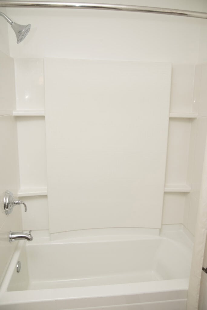 A clean, white bathtub and shower combination with a wall-mounted showerhead, faucet, and built-in shelves on both sides, all surrounded by a white shower wall panel.