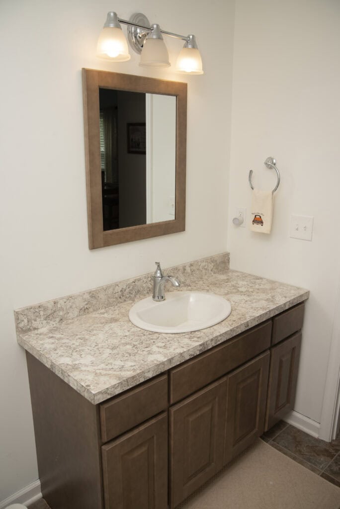 A bathroom vanity with a marble-patterned countertop, built-in sink, chrome faucet, and wooden cabinets. Above the sink is a framed mirror with two wall-mounted lights. A towel ring and light switches are on the wall.