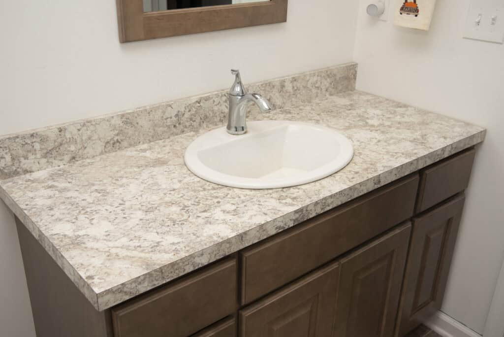A bathroom vanity with a light-colored, marbled countertop, a round white sink, a single-handle faucet, and wooden cabinets below. A mirror and towel holder are partially visible on the white wall above.