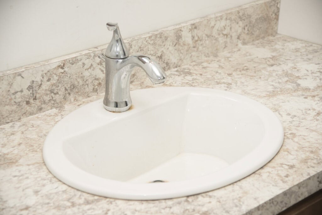 A white bathroom sink with a silver faucet is installed in a light-colored, marbled countertop. The sink area is clean and simple, with no visible accessories or decorations.