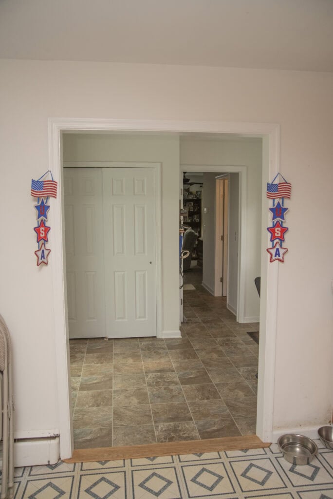A doorway decorated with USA signs and American flags leads to a room with tiled flooring. Sliding closet doors are on the left, and a glimpse of another room is visible through the open doorway. Dog bowls are on the floor.