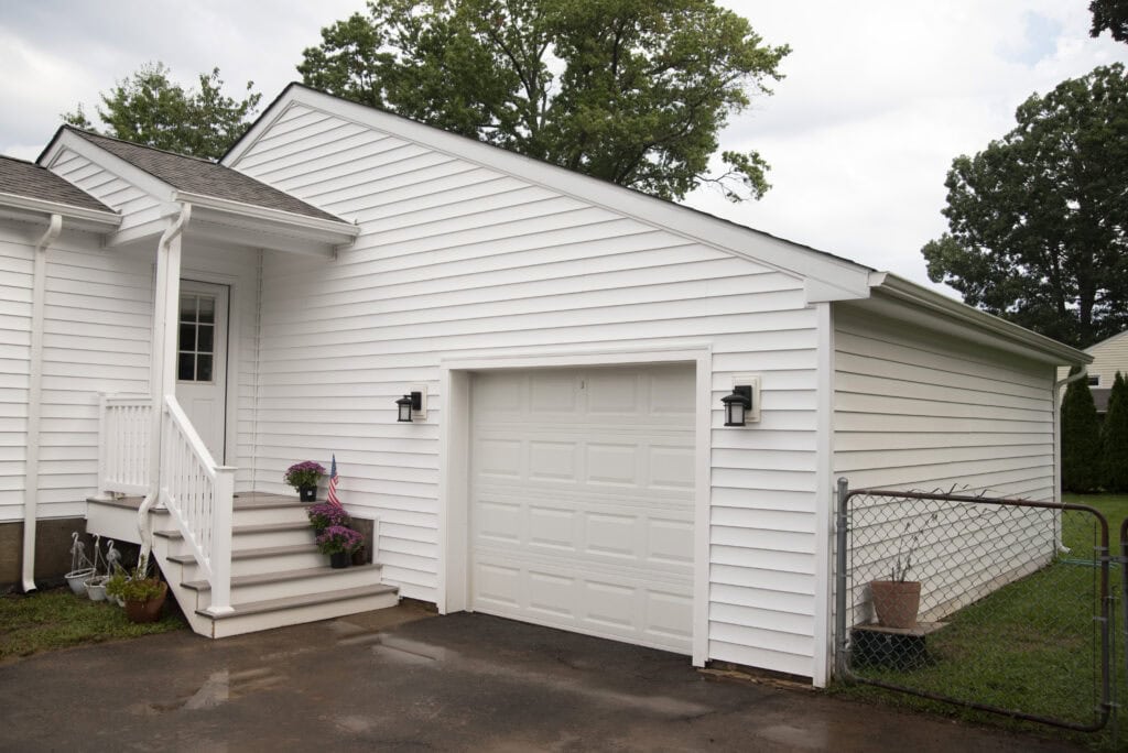A white, single-story house with white siding, a one-car garage, a small set of steps with a railing, potted plants on the steps, and a fenced yard. Trees are visible in the background.