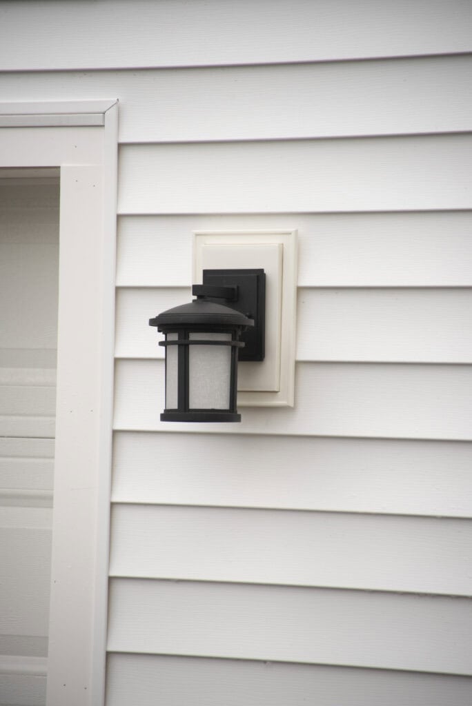 A black outdoor wall lantern with frosted glass is mounted on the white siding of a house near a doorframe.