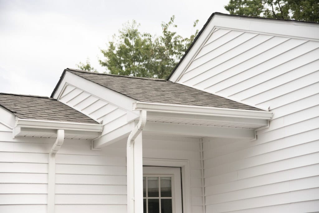 Close-up of the exterior corner of a white house with vinyl siding, showing a small overhang roof, white gutters, and a door beneath the roof. Trees and cloudy sky are visible in the background.