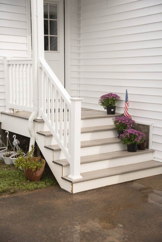 White house porch steps with three potted purple flowers and a small American flag on the steps. A potted plant and garden statue are beside the steps on the ground. The porch has a white railing.