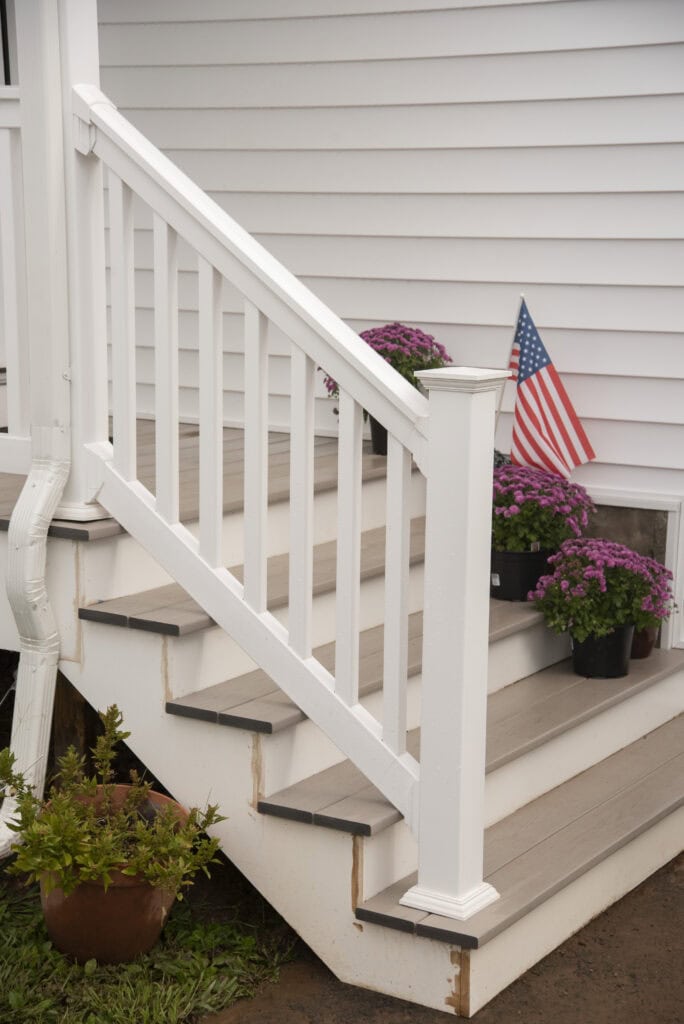 White wooden steps with a railing lead to a house with white siding. Purple potted flowers and a small American flag are placed on the steps. A brown flowerpot with greenery sits at the bottom.