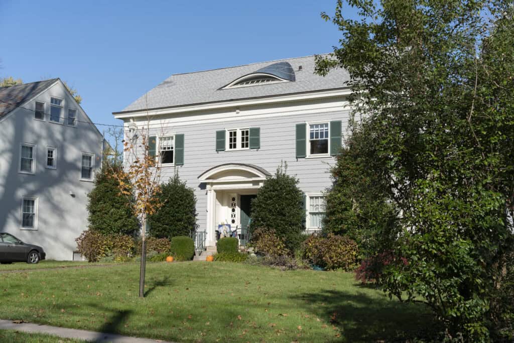 A two-story white house with green shutters, surrounded by bushes and trees, sits on a grassy lawn under a clear blue sky. Pumpkins decorate the front porch, and a gray car is parked beside a neighboring house.