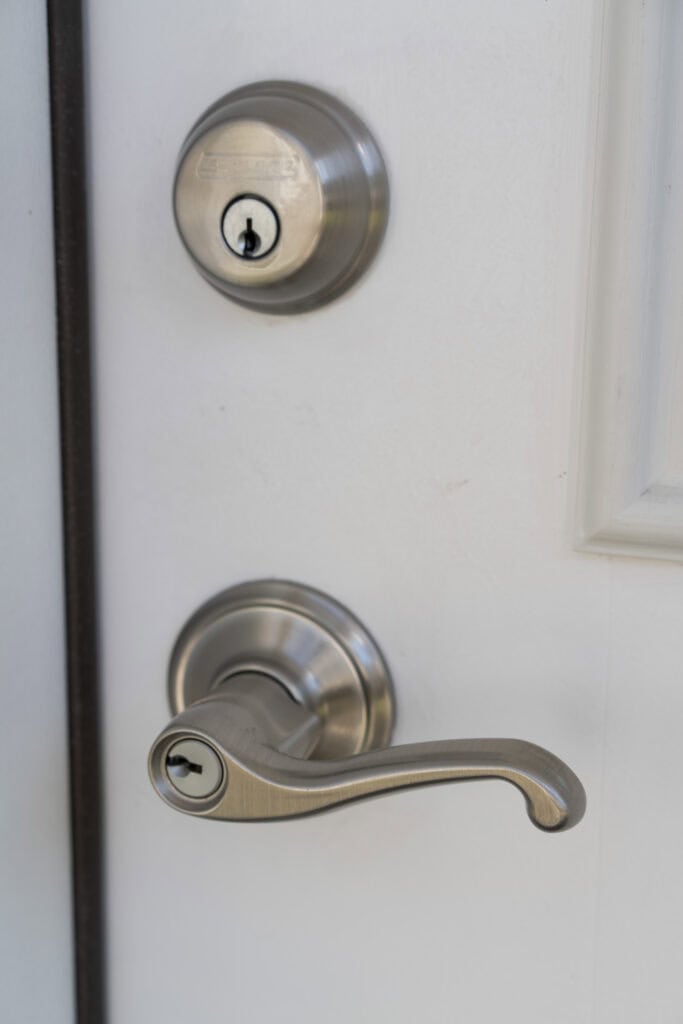 A close-up of a white door showing a silver deadbolt lock above a matching silver door handle with a keyhole.