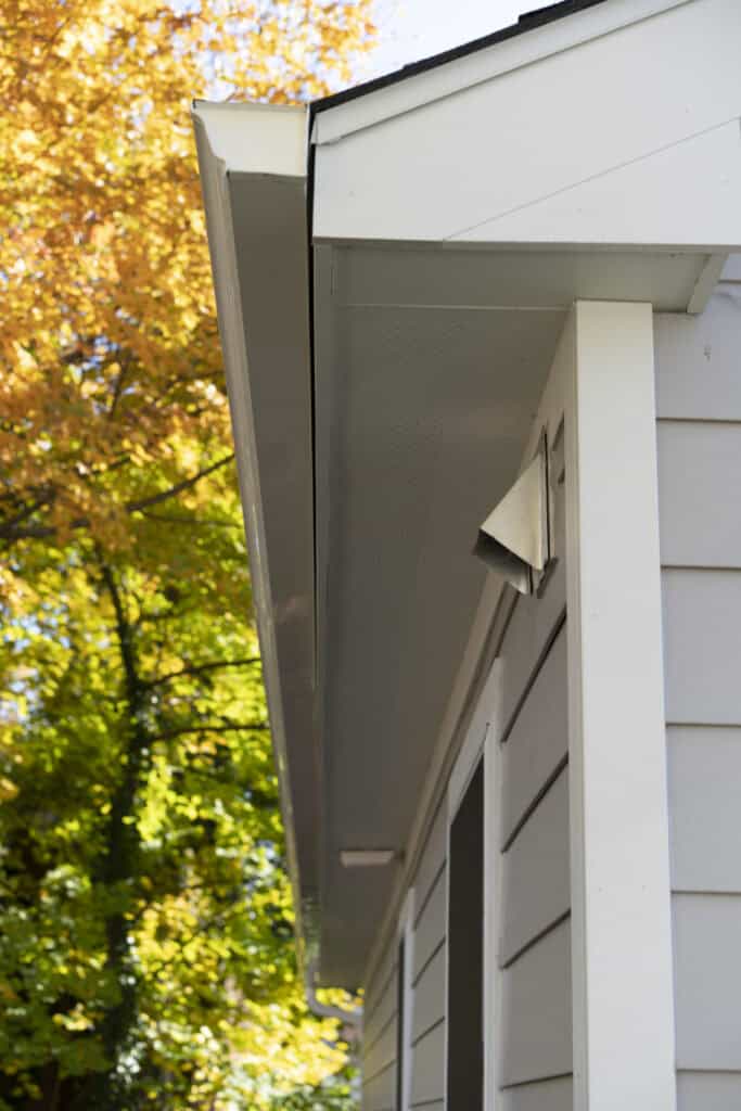 Close-up view of a house’s exterior, focusing on the white soffit, gutter, and siding. Yellow and green autumn trees are visible in the background under a clear sky.