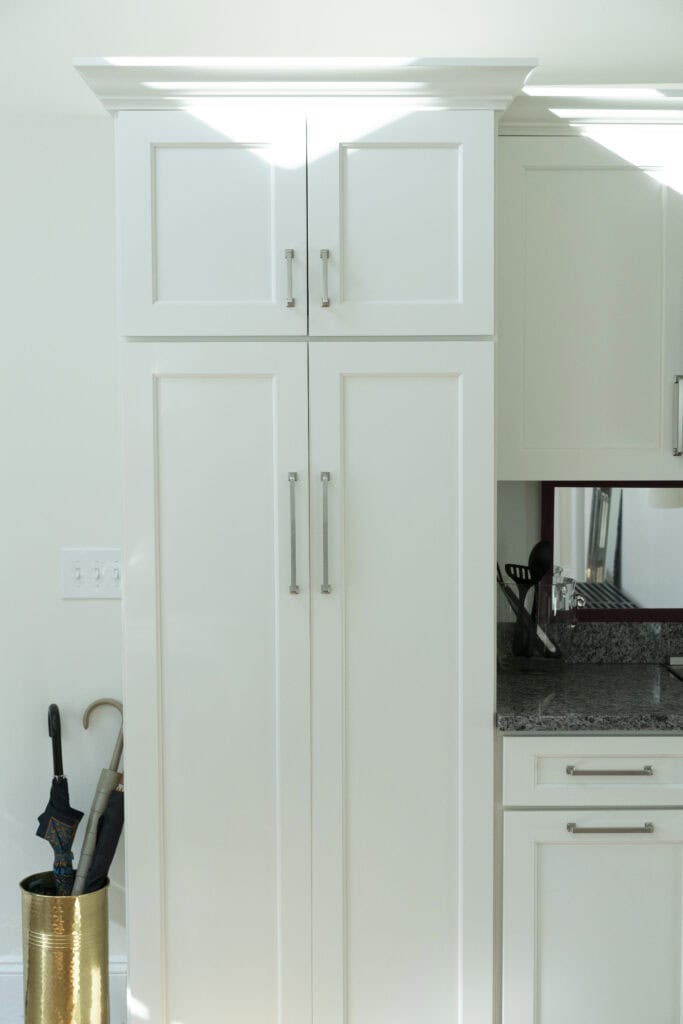 White kitchen cabinets with silver handles stand next to a granite countertop. On the left, a gold umbrella holder contains two umbrellas. Sunlight streams in, casting shadows on the cabinets and wall.