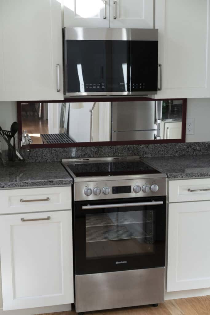 A modern kitchen with a stainless steel oven and stove, a black microwave above it, white cabinets, a granite countertop, and kitchen utensils on the left. A mirror backsplash reflects a refrigerator and floor.