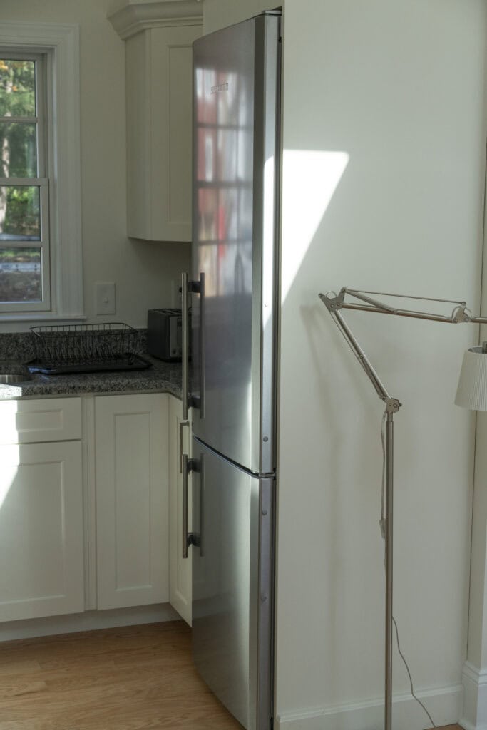 A stainless steel refrigerator stands in a corner of a white kitchen near cabinets and a granite countertop, with a floor lamp positioned close to it. Sunlight streams through a window, casting a shadow on the fridge.