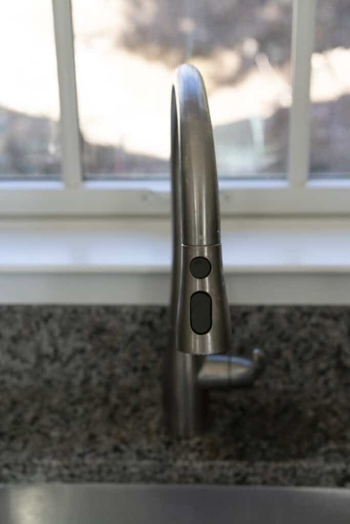 A modern stainless steel kitchen faucet with a curved spout and spray buttons, set in a granite countertop in front of a window with white framing.