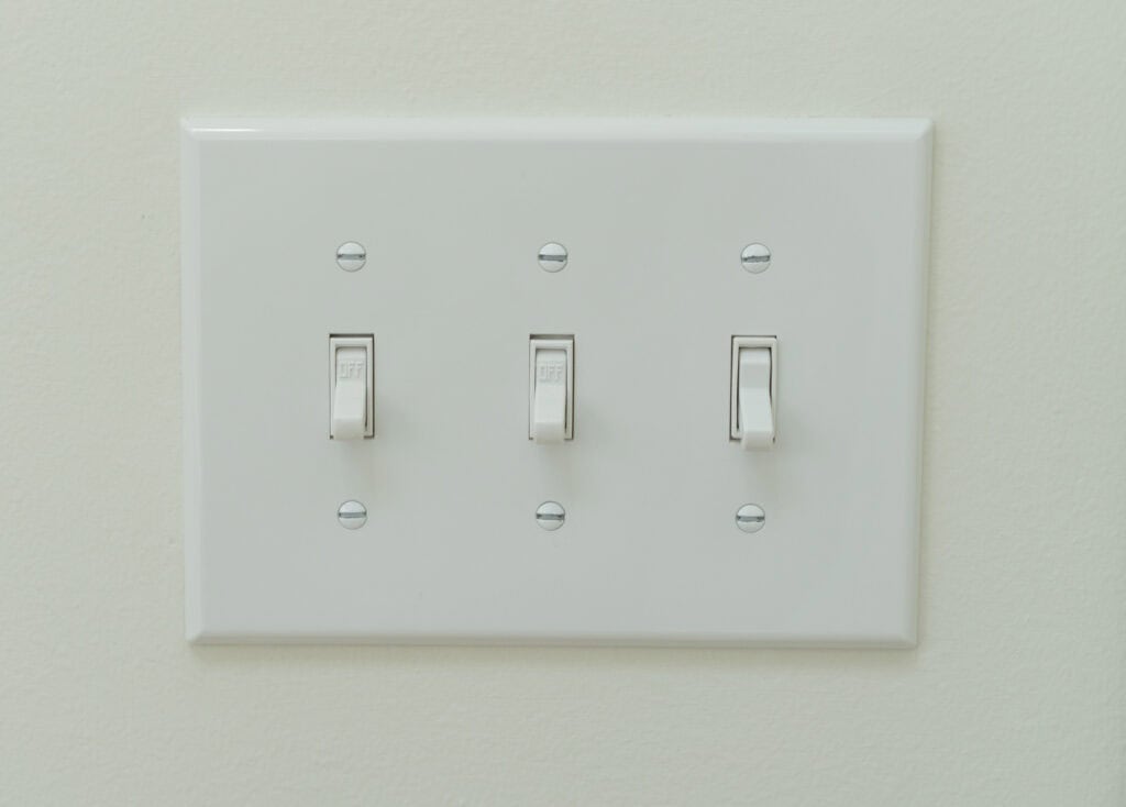 A white wall plate with three light switches; the left and center switches are in the off position, and the right switch is in the on position, against a pale wall.