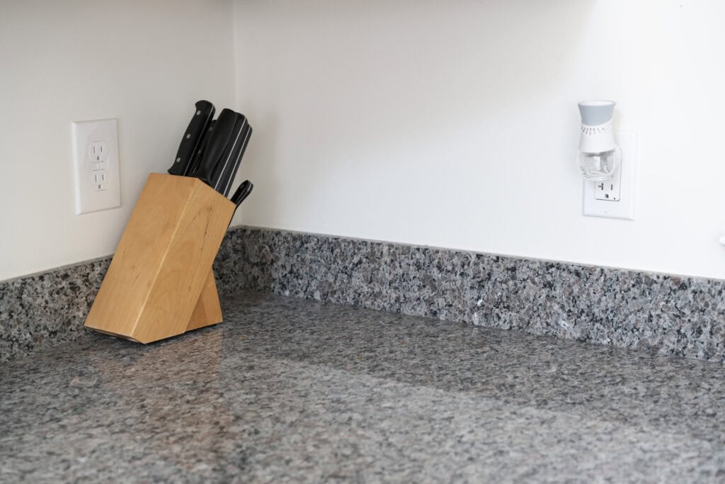 A wooden knife block with several knives sits on a gray granite kitchen countertop next to a wall outlet, with a white air freshener plugged into another outlet on the right.