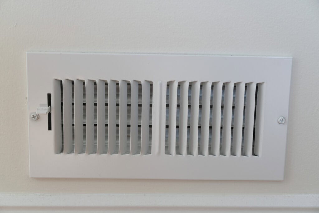 A white metal air vent cover is mounted on a light-colored wall, with horizontal and vertical slats and a small adjustable lever on the left side.