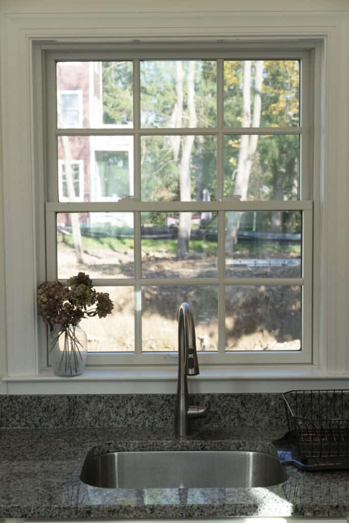A kitchen sink with a silver faucet is set in a granite countertop beneath a window. A vase with dried flowers sits on the left, and a drying rack is on the right. Sunlight streams through the window, showing trees outside.