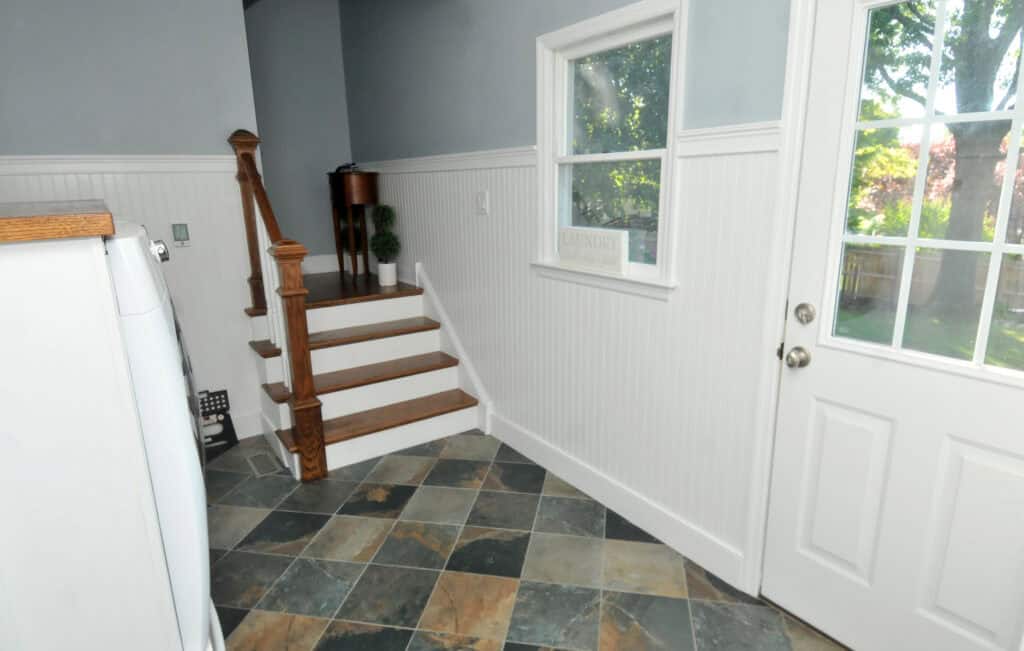 A laundry room with a tiled floor, white wainscoting, a window, and a door. Wooden stairs with a banister lead up to another area, and a small table with a plant is on the landing.