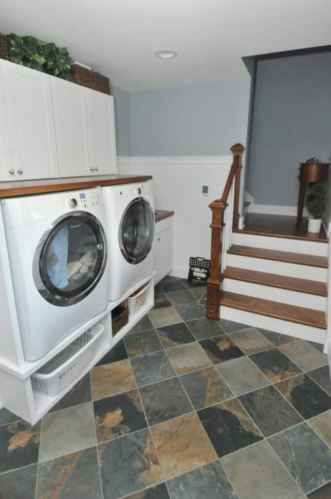 A laundry room with a front-loading washer and dryer set in white cabinetry, baskets below, slate tile flooring, white cabinets above, and wooden stairs leading up to a gray wall.
