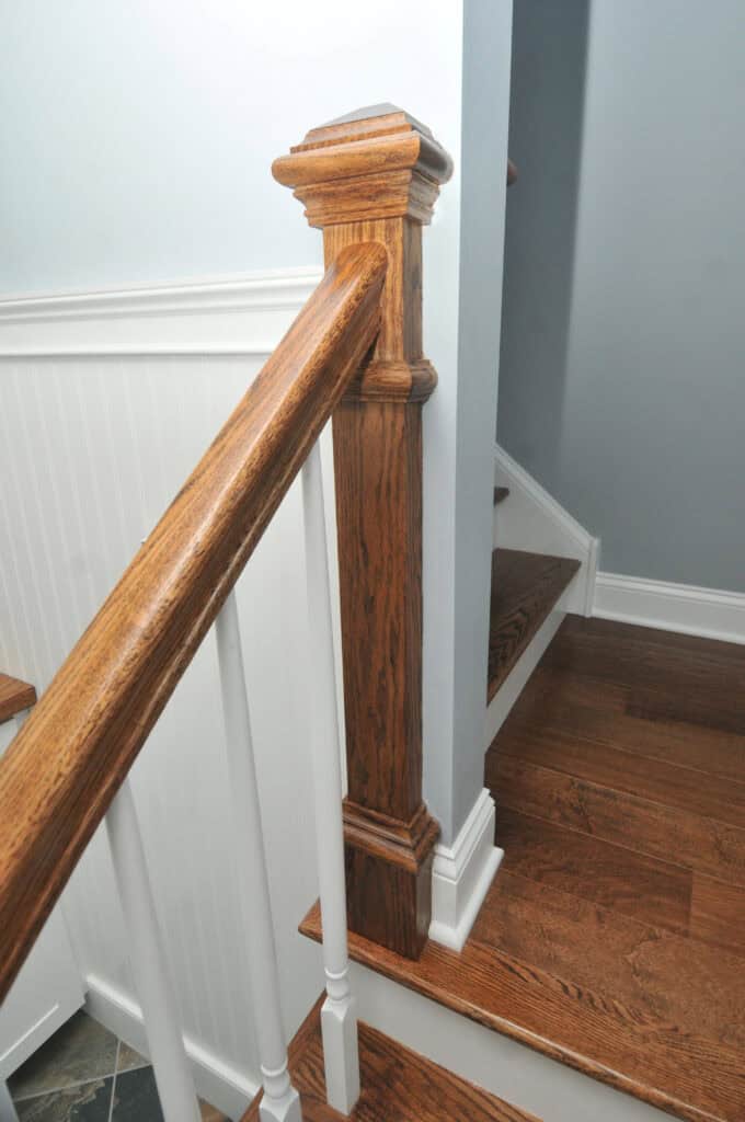A wooden staircase with oak handrail and newel post, white spindles, and white paneling on the lower wall, next to light blue painted walls.