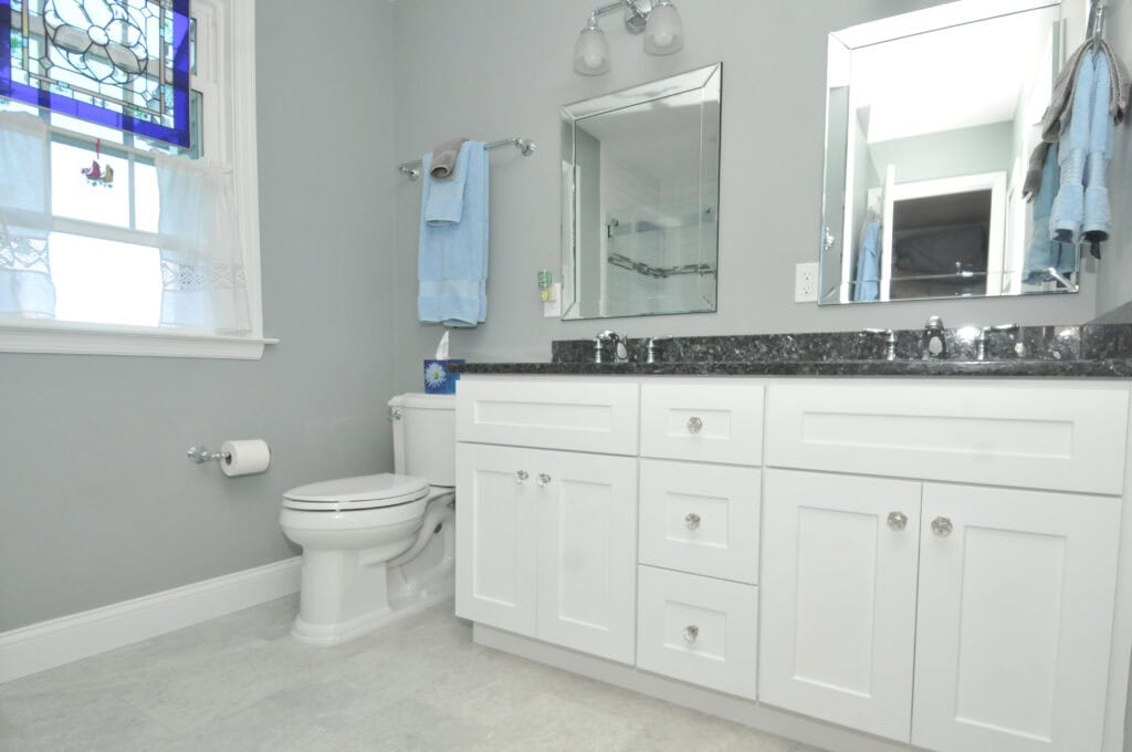 A clean, modern bathroom with white cabinets, a black countertop, a toilet, a towel rack, and a window with a stained glass design. There are two mirrors and soft gray walls.