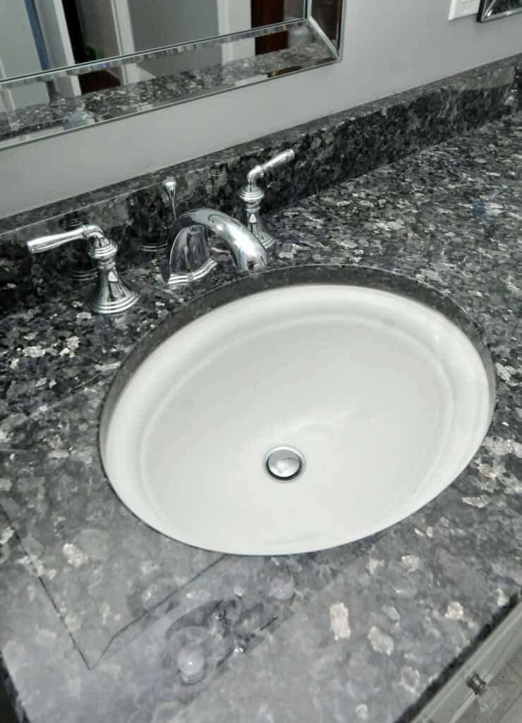 A white oval sink set into a dark, speckled granite countertop with chrome faucet and handles; a mirror is visible above the sink.