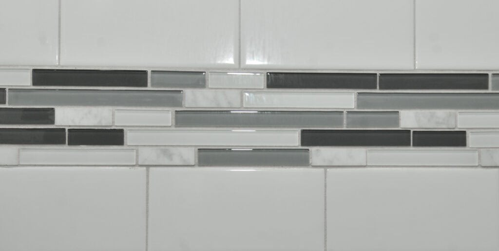 A close-up of a wall with white rectangular tiles and a horizontal accent strip of narrow glass tiles in shades of gray, white, and black.