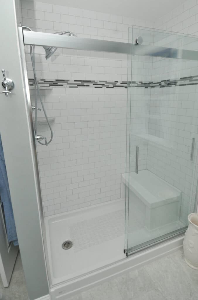 A modern shower with sliding glass doors, white subway tile walls, a built-in bench, and a silver showerhead. There is a decorative horizontal stripe of gray and black tiles on the wall.