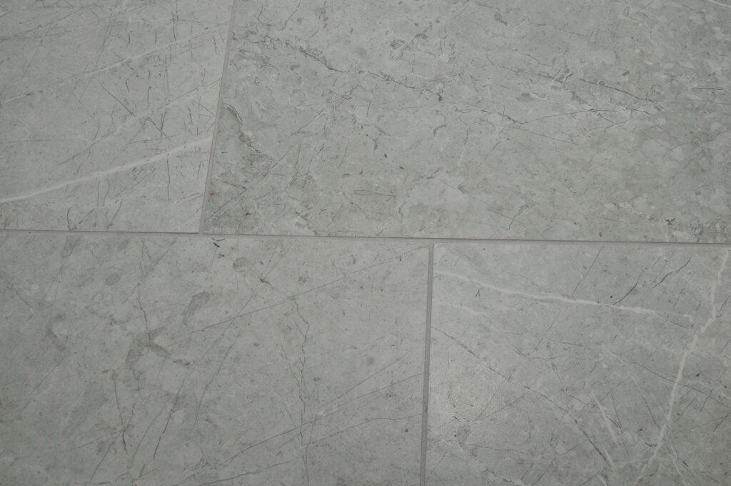 Close-up of light gray marble tiles with visible white and gray veining and natural cracks, showing the texture and pattern of the stone surface.