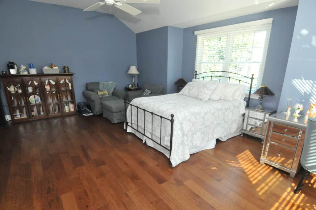A spacious bedroom with blue walls, a large window, a bed with a white and gray patterned comforter, an armchair, a wooden display cabinet, hardwood floors, and sunlight streaming onto the floor.