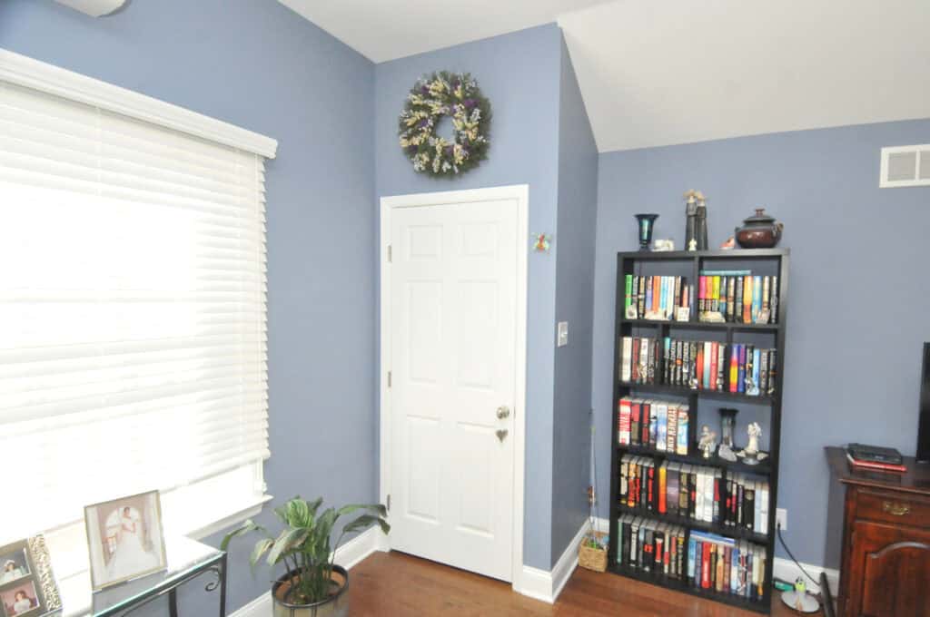 A white door with a floral wreath is set in a blue wall next to a window with blinds. Beside the door is a black bookshelf filled with books and decorative items. A small plant sits on the wood floor.