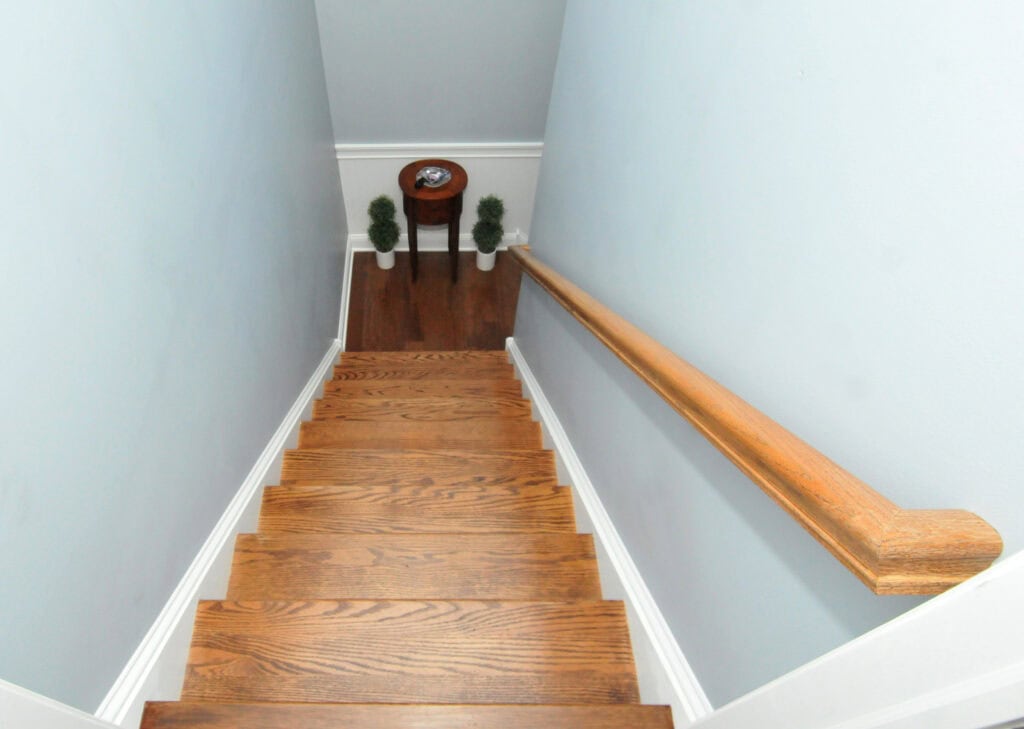 A view looking down a wooden staircase with a brown handrail on the right. At the bottom, a small round table with a dish and two potted plants on either side sits against a light blue wall.