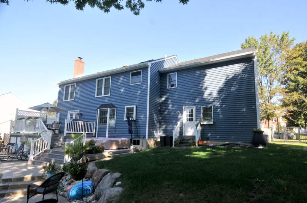 A two-story blue house with white trim, multiple windows, and a backyard patio. The yard features green grass, landscaping with rocks, and outdoor furniture under a clear, sunny sky.