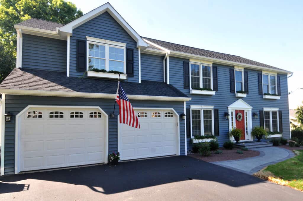 A two-story blue house with white trim, black shutters, and a red front door. There are three garage doors, an American flag, and a paved driveway and walkway bordered by small shrubs and plants.