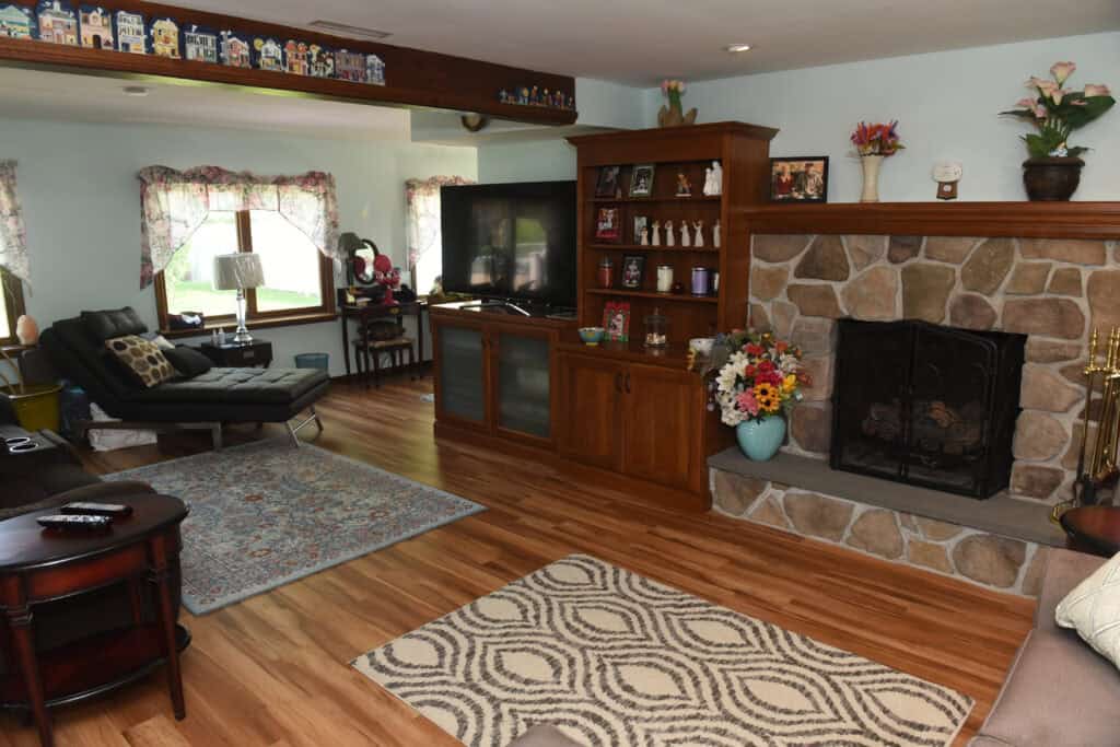 A cozy living room with hardwood floors, patterned rugs, a stone fireplace, a TV on a wooden console, shelves with photos and figurines, a lounge chair, and floral curtains on the windows.