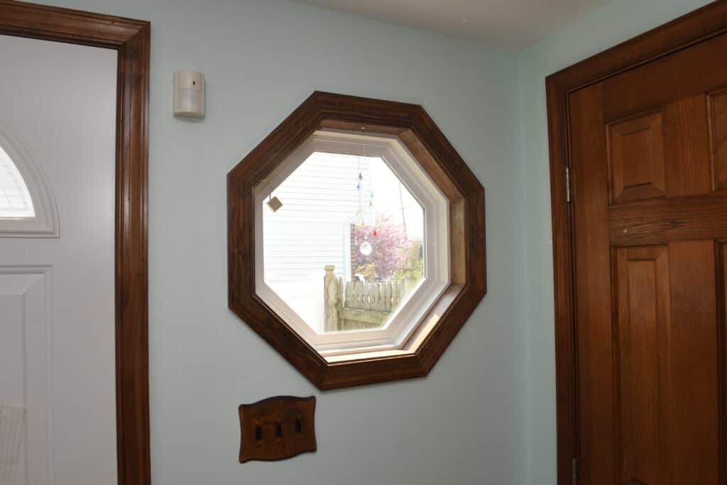 A wooden octagon-shaped window with a view of a yard and plants, set in a light-colored wall between a white door and a wooden door inside a house.