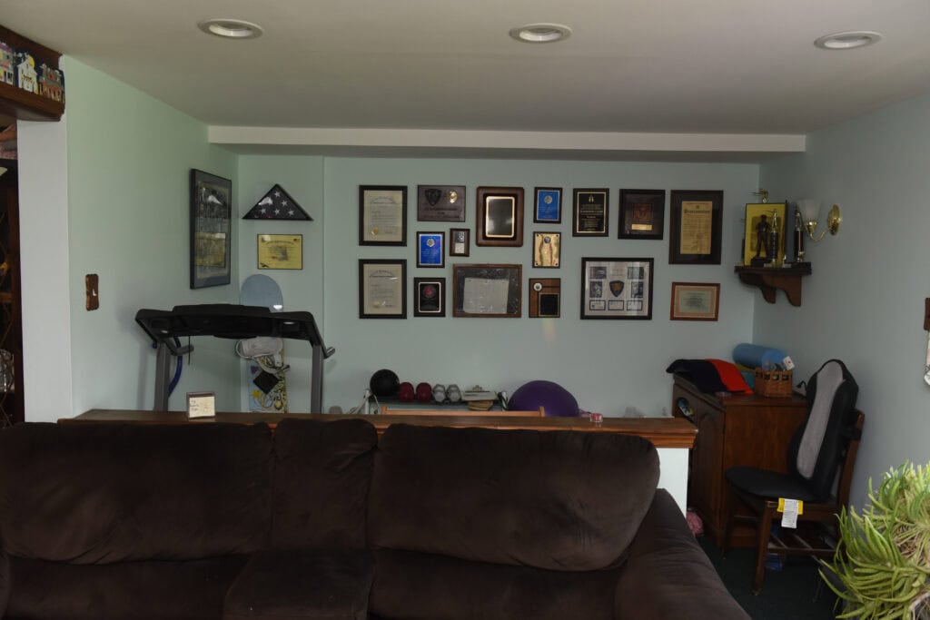 A room with a dark brown couch in the foreground, a treadmill, and a collection of plaques, awards, and framed certificates on a light green wall. Theres also a desk with various items and exercise equipment visible.