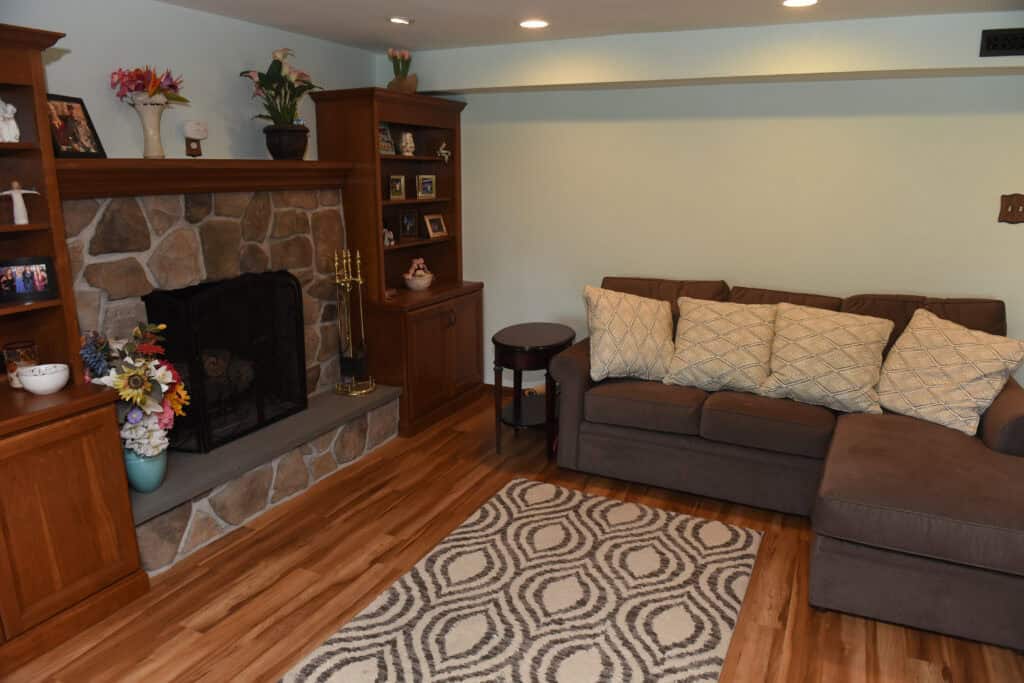 A cozy living room with a stone fireplace, wooden shelves with photos and decor, a gray sectional sofa with patterned pillows, a round side table, and a geometric-patterned rug on a wooden floor.