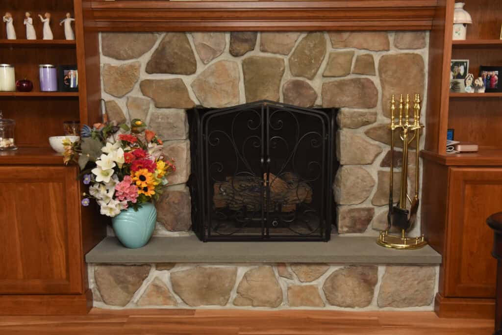 A stone fireplace with a black metal screen, flanked by a blue vase of colorful artificial flowers on the left and brass fireplace tools on the right. Wooden shelves and cabinets surround the fireplace.