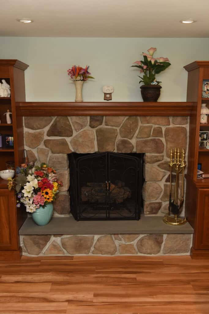 A stone fireplace with a black metal screen, flanked by wooden shelves. Floral arrangements and plants decorate the mantle, while brass fireplace tools sit on the right side. Wooden flooring is visible in front.