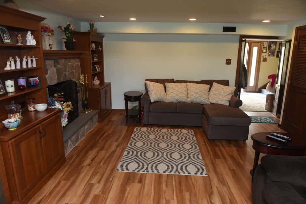 A cozy living room with wood flooring, a patterned rug, a brown sectional sofa with cushions, wooden shelves, a stone fireplace, and decorative items. There is natural light coming from an adjacent hallway.