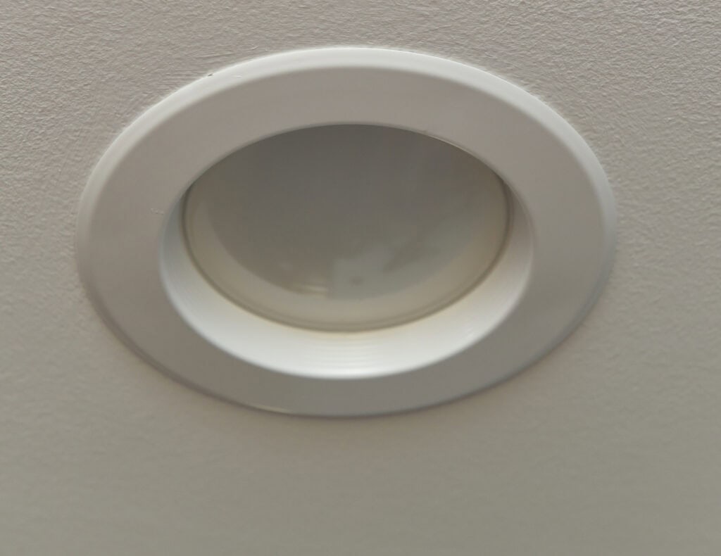 A close-up photo of a recessed ceiling light fixture with a white trim, set into a light-colored textured ceiling. The light is turned off.