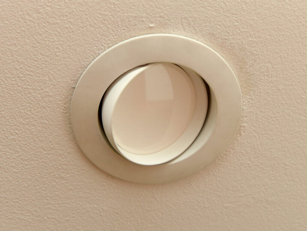 A close-up of a round recessed ceiling light with a white frame, mounted in a textured beige ceiling. The light is off, and the glass cover is clean and slightly reflective.
