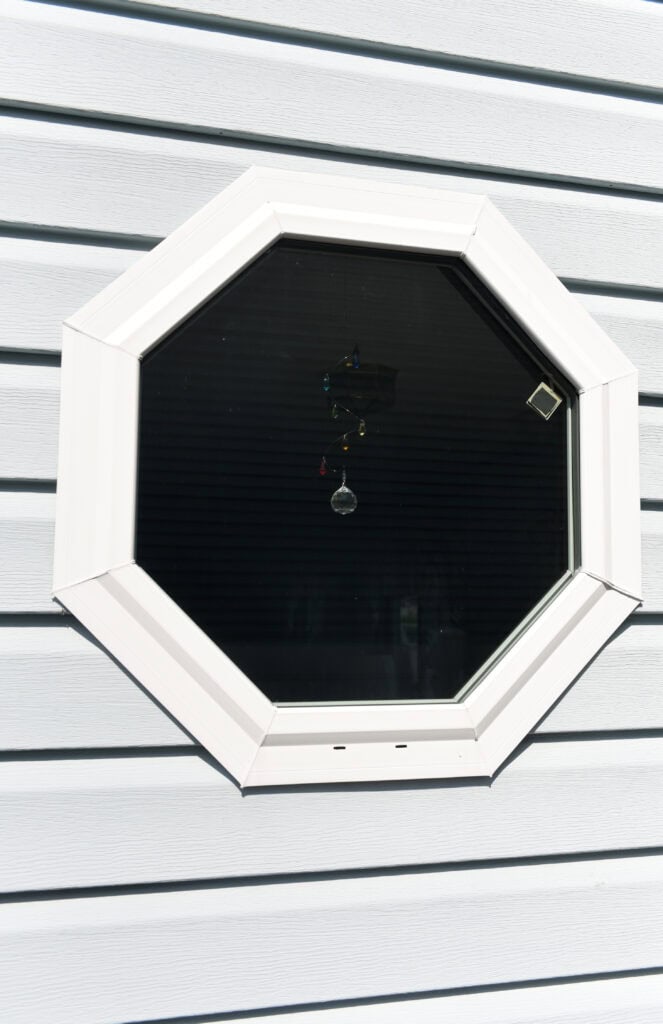An octagonal window with a white frame set in light gray siding. A small hanging crystal decoration is visible inside the window, reflecting light.