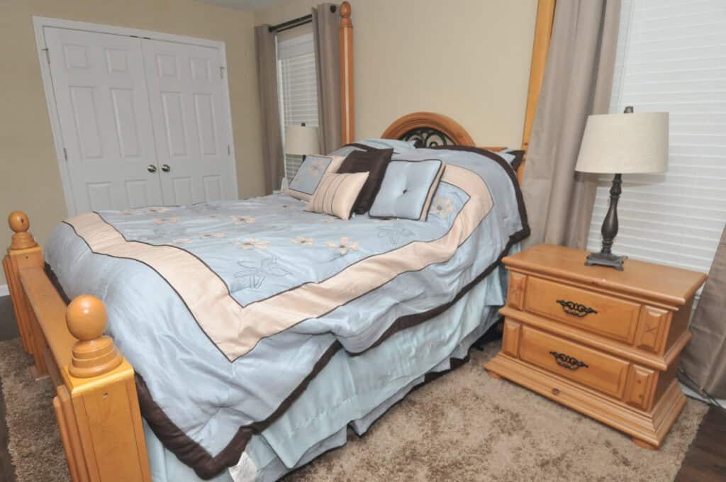 A neatly made bed with blue and beige floral bedding, several decorative pillows, and a wooden headboard. Beside the bed is a wooden nightstand with a lamp, in a carpeted bedroom with white doors and windows.